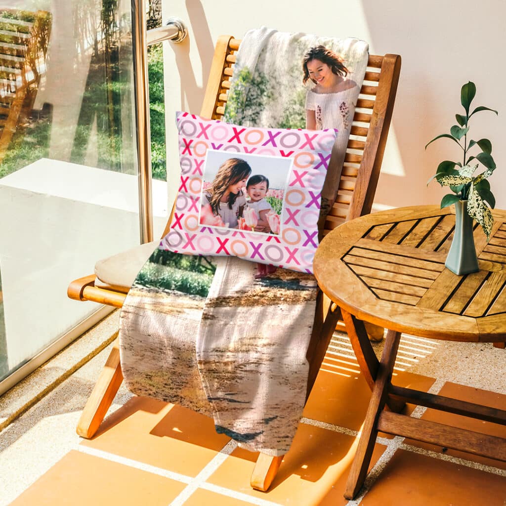 Personalised pillow and blanket on a deck chair beside a table with plant in a vase on it.